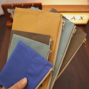 May include: A collection of colorful zippered wallets in shades of blue, green, brown, and yellow. Each wallet has a contrasting zipper and is made of a textured fabric. The wallets are fanned out, showcasing their different colors and designs.
