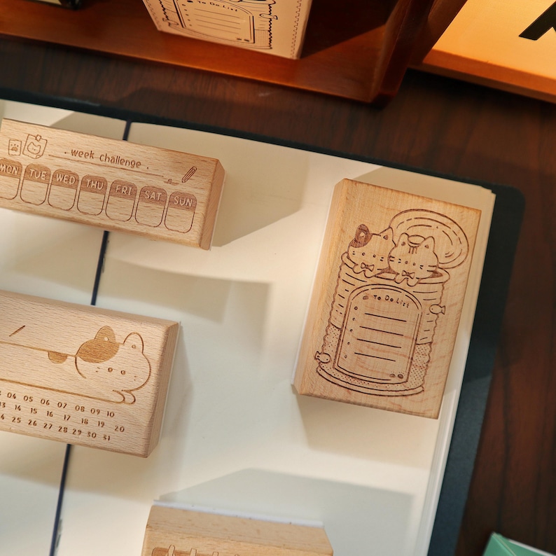 May include: Wooden stamps with various designs, including a weekly planner, a cat in a can, and a cat with a calendar. The stamps are made of light-colored wood and feature detailed illustrations and text. The stamps are designed for crafting and planning.