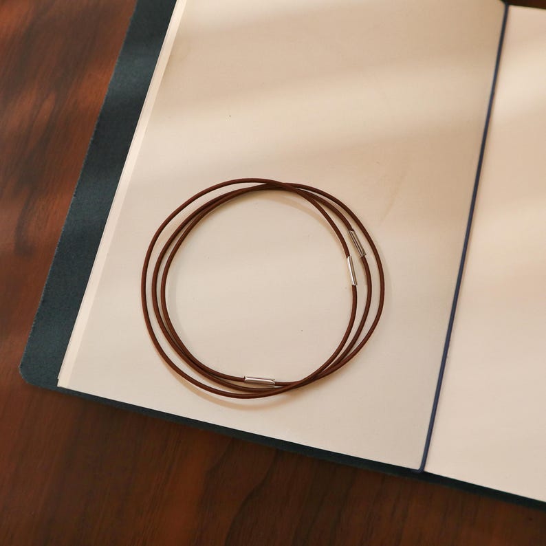 Pu&ograve; includere: Tre cordoncini in pelle marrone con chiusure in metallo argentato, disposti a cerchio, poggiano sulle pagine aperte di un taccuino. Il taccuino ha una copertina blu scuro e pagine interne bianche. I cordoncini sembrano essere per una collana o un braccialetto.