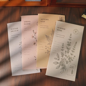 May include: Four rectangular paper notebooks featuring botanical illustrations and text. The covers are labelled Winter Branch, Spring Bloom, Autumn Leaf, and Summer Herb, each with unique plant drawings and printed phrases in a muted colour palette.