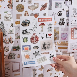 May include: A sticker sheet featuring various illustrations including a cat in a party hat, a mushroom, a pomegranate, black high-top sneakers, a vinyl record, a sun, stars, eyes, dancing cats, and text labels like "I love music" and "old soul."