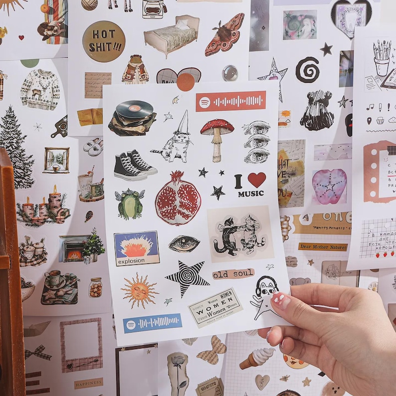 May include: A sticker sheet featuring various illustrations including a cat in a party hat, a mushroom, a pomegranate, black high-top sneakers, a vinyl record, a sun, stars, eyes, dancing cats, and text labels like "I love music" and "old soul."
