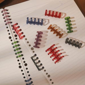Puede incluir: Una colección de coloridos anillos de encuadernación de plástico en rojo, azul, verde, marrón, morado y negro. Los anillos están dispersos en una página de cuaderno rayada, mostrando sus diversos diseños y colores. Ideales para organizar documentos.