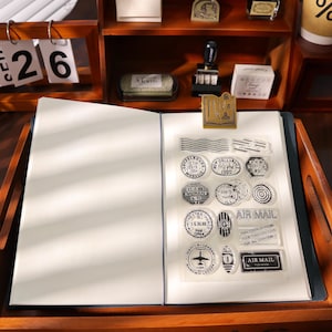 May include: An open notebook with blank pages, a clear sheet of stamps, and a gold-colored clip. The stamps feature various circular and rectangular designs with text, including "AIR MAIL" and "MANILA-LONDON". The scene is set on a wooden desk with a calendar.