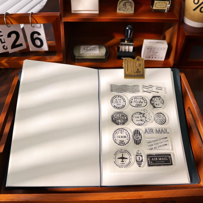 May include: An open notebook with blank pages, a clear sheet of stamps, and a gold-colored clip. The stamps feature various circular and rectangular designs with text, including "AIR MAIL" and "MANILA-LONDON". The scene is set on a wooden desk with a calendar.
