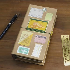 Peut inclure: Un carnet de voyage marron avec divers autocollants, dont une étiquette "VISIT PASS" et un timbre "SUMMER time". Un stylo noir repose sur la surface en bois à côté du carnet. Le carnet est maintenu par un élastique vert.