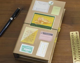 Op de afbeelding: Een bruin Traveler's notitieboek met diverse stickers, waaronder een "VISIT PASS" label en een "SUMMER time" stempel. Een zwarte pen ligt op het houten oppervlak naast het notitieboek. Het notitieboek wordt bij elkaar gehouden door een groene elastiek.