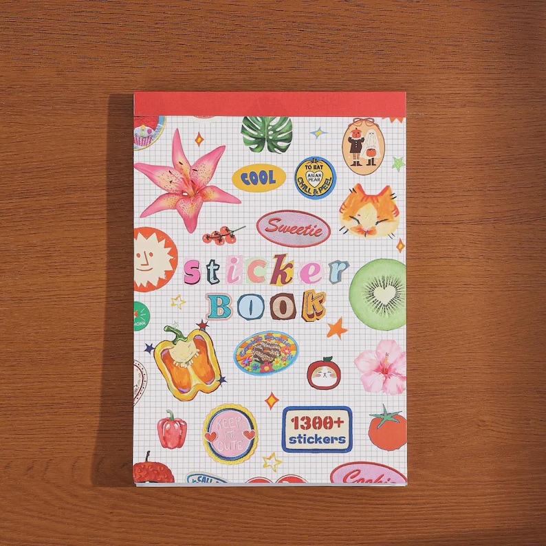 May include: A rectangular sticker book with a red top binding and a white grid background. The cover features colorful illustrations of a pink lily, a kiwi slice, a bell pepper, a tomato, a cat face, and various text labels including "sticker BOOK" and "1300+ stickers".