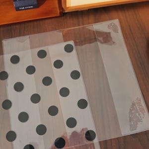 May include: Clear plastic sheets, one with a pattern of black polka dots. The sheets are partially overlapping, with the polka dot sheet on the bottom. The text "PAR AVION" is visible on a small blue box in the background.