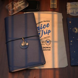 Op de afbeelding: Een marineblauw leren notitieboek met een fietsbedel, een cr&egrave;mekleurige reistas met de tekst "Nice Trip" en andere schrijfwaren. Het notitieboek is afgesloten met een elastiek.
