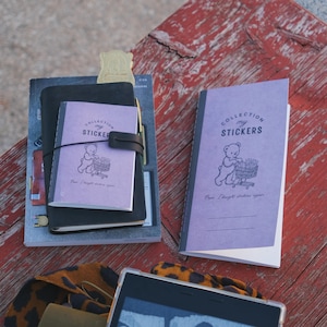 May include: Two light purple notebooks featuring a graphic of a bear pushing a shopping cart filled with stickers. The text reads "COLLECTION my STICKERS" and "Oops I bought stickers again." One notebook is placed on a black leather cover.