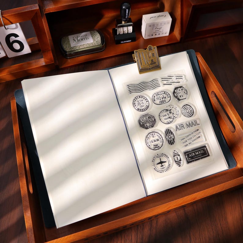 May include: A clear sheet of stamps with various vintage designs, including circles, rectangles, and wavy lines. The stamps feature text like "AIR MAIL" and city names. The sheet is on a notebook with blank pages, inside a wooden tray.