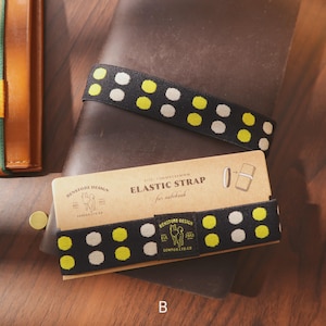 May include: Black elastic strap with yellow and white polka dots, designed for notebooks. The strap is displayed on a brown leather notebook with a matching cardboard sleeve. The sleeve features the text "Elastic Strap for notebook" and the brand name "Benstone Design."