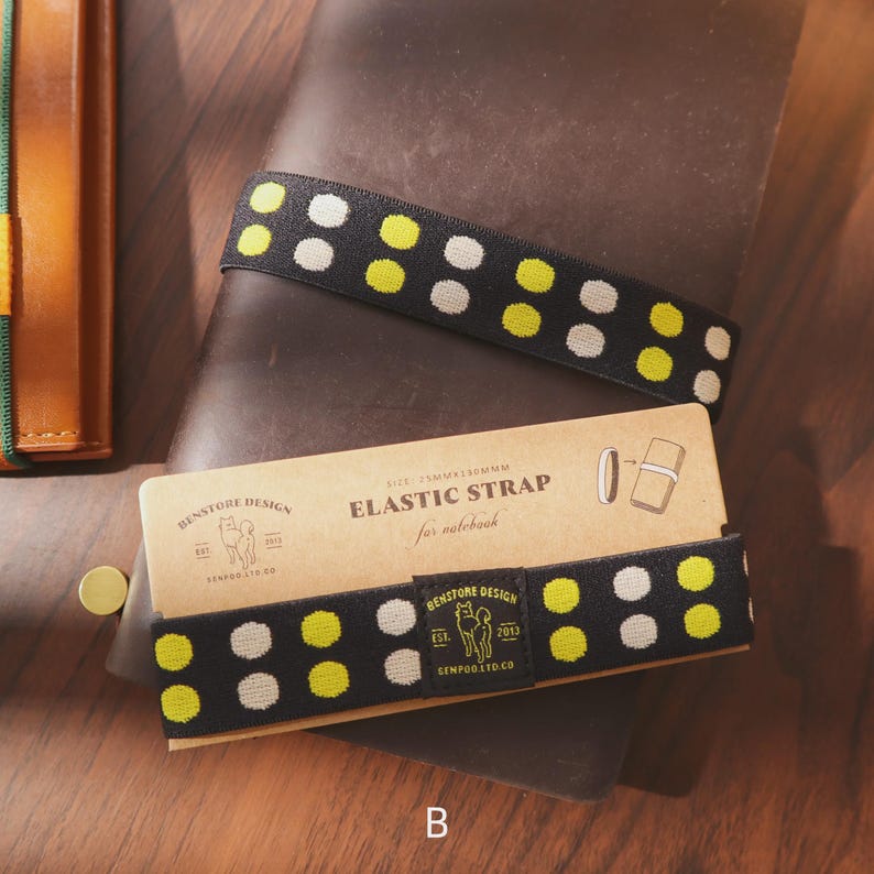 May include: Black elastic strap with yellow and white polka dots, designed for notebooks. The strap is displayed on a brown leather notebook with a matching cardboard sleeve. The sleeve features the text "Elastic Strap for notebook" and the brand name "Benstone Design."
