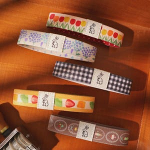 May include: An assortment of decorative fabric bands with various patterns, including floral, gingham, and fruit designs. Each band features a small white label with a rabbit illustration. The bands are arranged on a wooden surface.