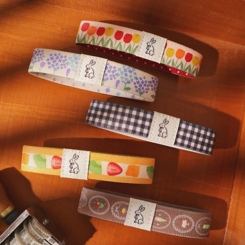 May include: An assortment of decorative fabric bands with various patterns, including floral, gingham, and fruit designs. Each band features a small white label with a rabbit illustration. The bands are arranged on a wooden surface.
