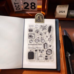 May include: A clear stamp set featuring a camera, bicycle, and film strip designs is shown on an open notebook. A metal bookmark with a cat and moon design is clipped to the notebook. The words "Thank You" are also visible.