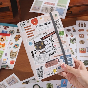 May include: A light-colored notebook covered in various stickers, including a red heart button, a star-patterned mug, a treble clef, and text phrases like "Think Of Me" and "Witchy Vibes." A dark gray elastic band secures the notebook closed.