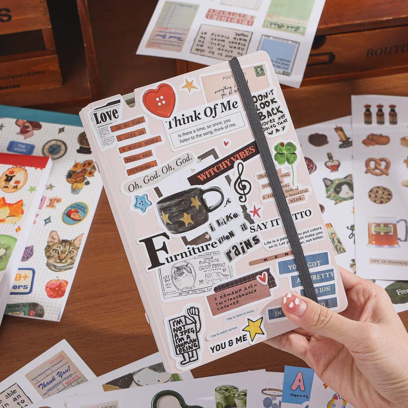 May include: A light-colored notebook covered in various stickers, including a red heart button, a star-patterned mug, a treble clef, and text phrases like "Think Of Me" and "Witchy Vibes." A dark gray elastic band secures the notebook closed.