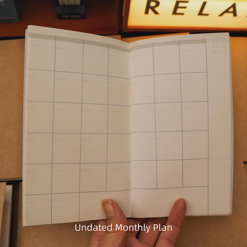 May include: An open, undated monthly plan notebook with a grid layout. The pages are white with gray grid lines. The text "Undated Monthly Plan" is at the bottom of the page.