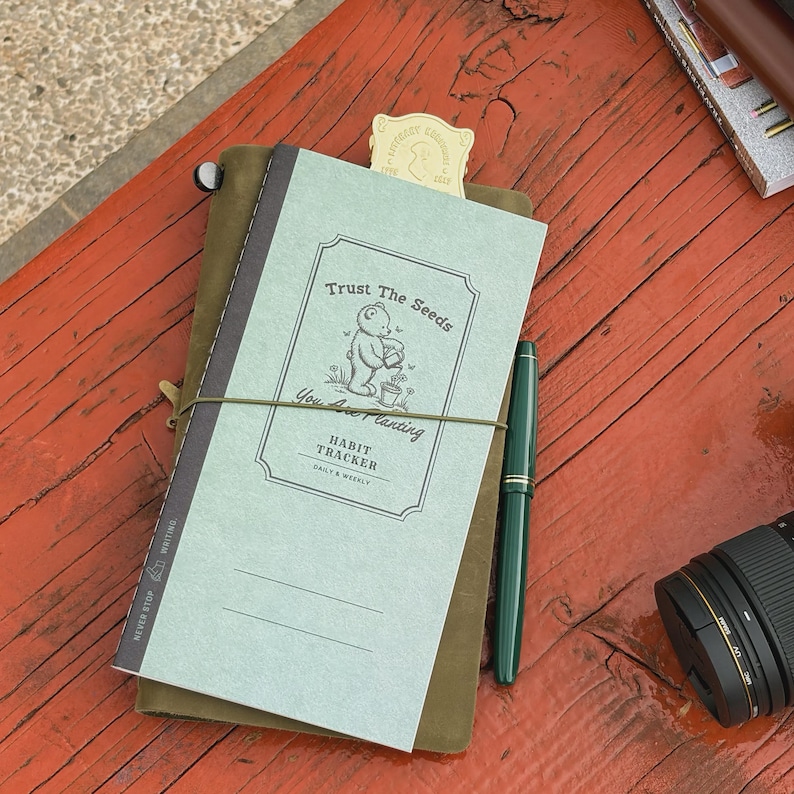 May include: A journal with the text "Trust The Seeds" and "You Are Planting" on the cover, along with a cartoon bear illustration. A green pen and a camera lens are also visible. The journal is bound with an elastic band.