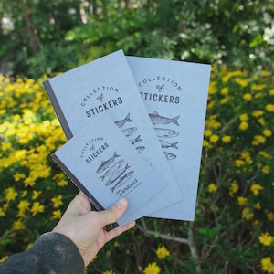May include: A collection of stickers with a fish theme. The light blue notebooks have the words "Collection of Stickers" and "Sardines" printed on them, along with illustrations of fish. The notebooks are held in front of a background of yellow flowers and green foliage.