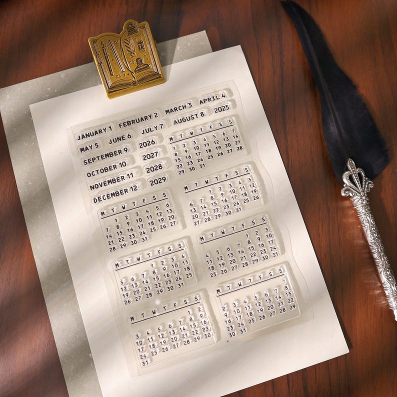 May include: A collection of clear acrylic calendar stamps with dates from 2025 to 2029, arranged on white paper. A gold-colored book-shaped embellishment and a quill pen are also present. The stamps are for planning and journaling.