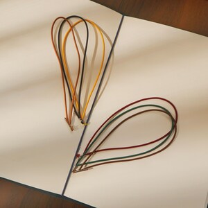 May include: A collection of colorful leather cords in various shades of brown, yellow, and red, arranged on a white surface. The cords are looped and appear to be for crafting or jewelry making. The image is well-lit, highlighting the textures and colors.