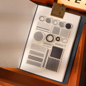 May include: A clear stamp set featuring diverse geometric designs such as circles, stripes, and grids. The stamps are displayed on a transparent sheet, ideal for scrapbooking and crafting. A gold-toned book clip secures the sheet within a notebook.