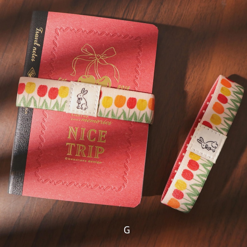 May include: A red notebook with gold lettering and a decorative elastic band featuring a tulip pattern and a small bunny illustration. The text "NICE TRIP" is visible on the notebook. A second band is also visible.