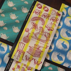 May include: Notebooks with various illustrated designs. One notebook features a yellow and white striped pattern with red and yellow illustrations of bears, cherries, and a rocking horse. Other notebooks have teal and blue patterns.