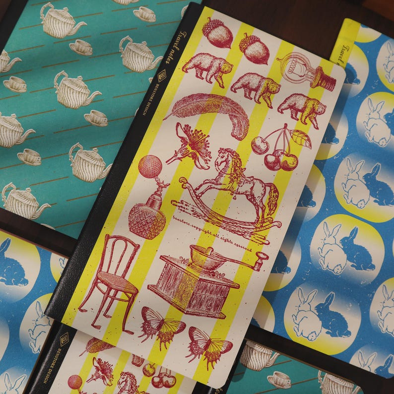 May include: Notebooks with various illustrated designs. One notebook features a yellow and white striped pattern with red and yellow illustrations of bears, cherries, and a rocking horse. Other notebooks have teal and blue patterns.