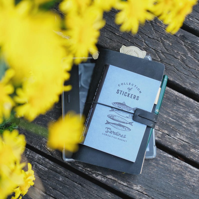 May include: A stack of notebooks and a pen rest on a wooden surface, with yellow flowers in the foreground. The top notebook is light blue with the words "Collection of Stickers" and illustrations of fish. The notebook is bound with a black elastic band.