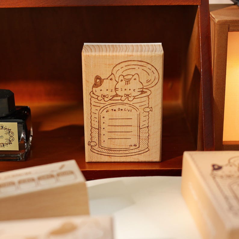 May include: A wooden stamp featuring a whimsical design of two cats in a can, with the words "To Do List" and lined spaces for notes. The stamp is made of light-colored wood and is ideal for stationery and crafting projects.