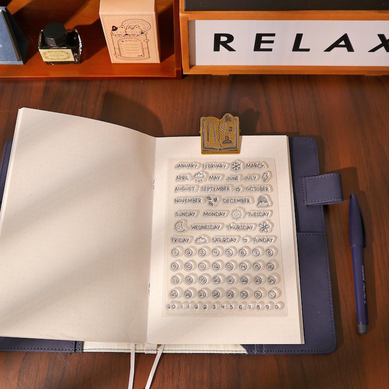 May include: An open notebook with a clear stamp sheet featuring months, days, and numbers. A gold-colored book-shaped paper clip holds the stamp sheet in place. A blue pen rests on the right side of the notebook. The word "RELAX" is visible in the background.