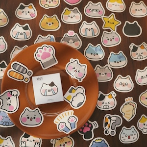 May include: A collection of cat-themed stickers with various expressions and designs. The stickers are in different colors, including yellow, gray, and blue. Some stickers feature paw prints and other cat-related graphics. A small notepad with a crying cat illustration is also present.