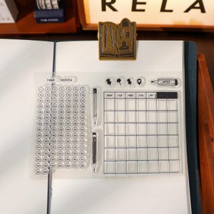 May include: A clear acrylic stamp set for planners and journals. Includes a habit tracker, monthly calendar, and various icons. A gold-coloured metal book-shaped paper clip is attached to the journal.