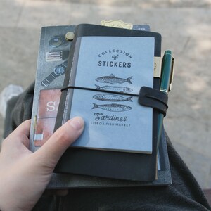 May include: A collection of notebooks and stickers featuring a blue cover with sardine illustrations and the text "Collection of Stickers" and "Sardines Lisbon Fish Market." A green pen is attached to the notebook with a black leather strap.