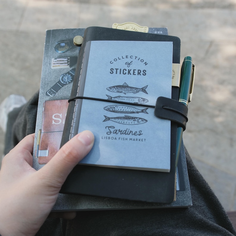 May include: A collection of notebooks and stickers featuring a blue cover with sardine illustrations and the text "Collection of Stickers" and "Sardines Lisbon Fish Market." A green pen is attached to the notebook with a black leather strap.