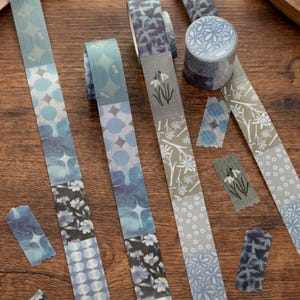 May include: Washi tape rolls and strips in blue, grey, and white. The tape displays floral, geometric, and abstract patterns. Some strips are torn and scattered on a wooden surface, with measurements in centimetres.