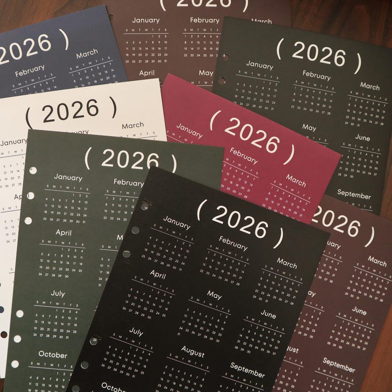 Pu&ograve; includere: Una collezione di pagine di calendario del 2026 in vari colori, tra cui nero, bordeaux, verde scuro e bianco. Ogni pagina mostra i mesi dell'anno, con l'anno 2026 in evidenza. Le pagine sono perforate per la rilegatura.