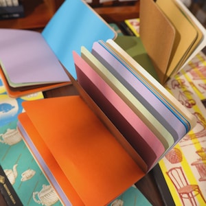 May include: Open notebooks with colorful pages. The notebooks have orange, blue, and purple covers. The pages are various pastel colors. The notebooks are on a table with other stationery items.