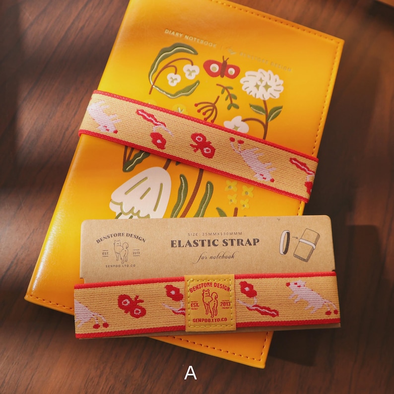 May include: A yellow diary notebook with floral illustrations and an elastic strap. The strap is tan with a red border and repeating floral and animal designs. A matching elastic strap and packaging are also visible.