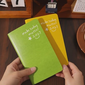 Könnte beinhalten: Zwei rechteckige Notizbücher, eines in Limettengrün und eines in Senfgelb, die jeweils den weißen Aufdruck "make today matter 2027" zusammen mit einer handgezeichneten Stern- und Smiley-Grafik auf dem Vorderdeckel tragen.