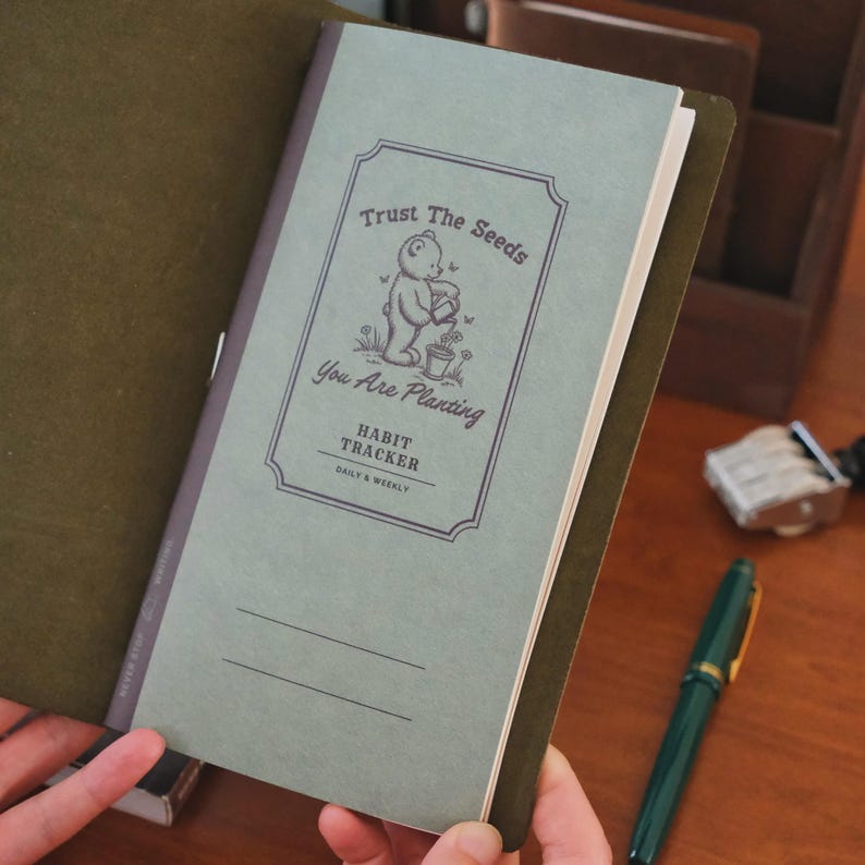 May include: A habit tracker journal with a sage green cover and a brown leather cover. The journal features the text "Trust The Seeds You Are Planting" with an illustration of a bear. The journal is open, revealing lined pages.
