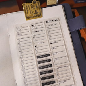 May include: A clear acrylic daily planner insert with a "Daily Plan" header, featuring sections for dates, goals, and a to-do list. The insert includes spaces for recording meals and expenses. A gold-colored book-shaped paper clip is attached to the planner.