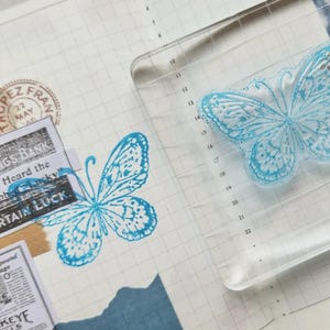 Puede incluir: Un sello de mariposa de acr&iacute;lico transparente con un dise&ntilde;o de mariposa azul se muestra junto a una imagen de mariposa estampada. El sello est&aacute; sobre un fondo de papel cuadriculado con recortes de peri&oacute;dicos y el texto "CERTAIN LUCK".