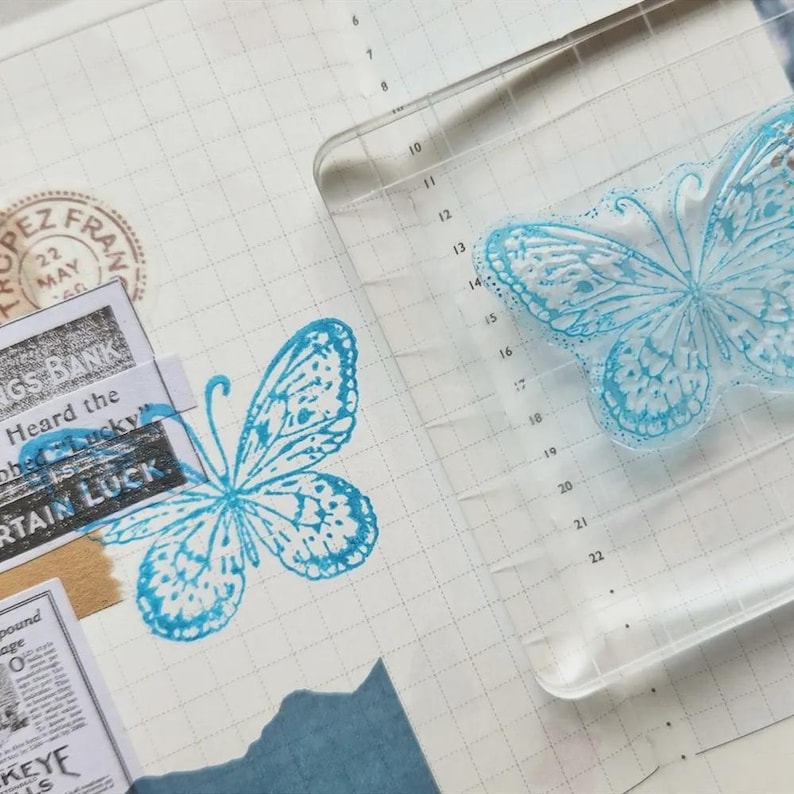 Puede incluir: Un sello de mariposa de acr&iacute;lico transparente con un dise&ntilde;o de mariposa azul se muestra junto a una imagen de mariposa estampada. El sello est&aacute; sobre un fondo de papel cuadriculado con recortes de peri&oacute;dicos y el texto "CERTAIN LUCK".