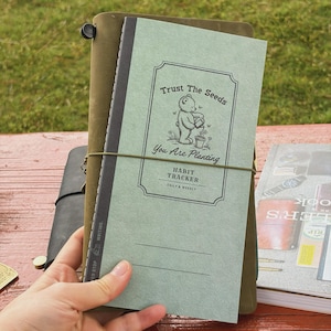 May include: A green habit tracker notebook with the text "Trust The Seeds" and "You Are Planting". The notebook is held together with an elastic band and is surrounded by other notebooks. The cover features a bear watering a plant.