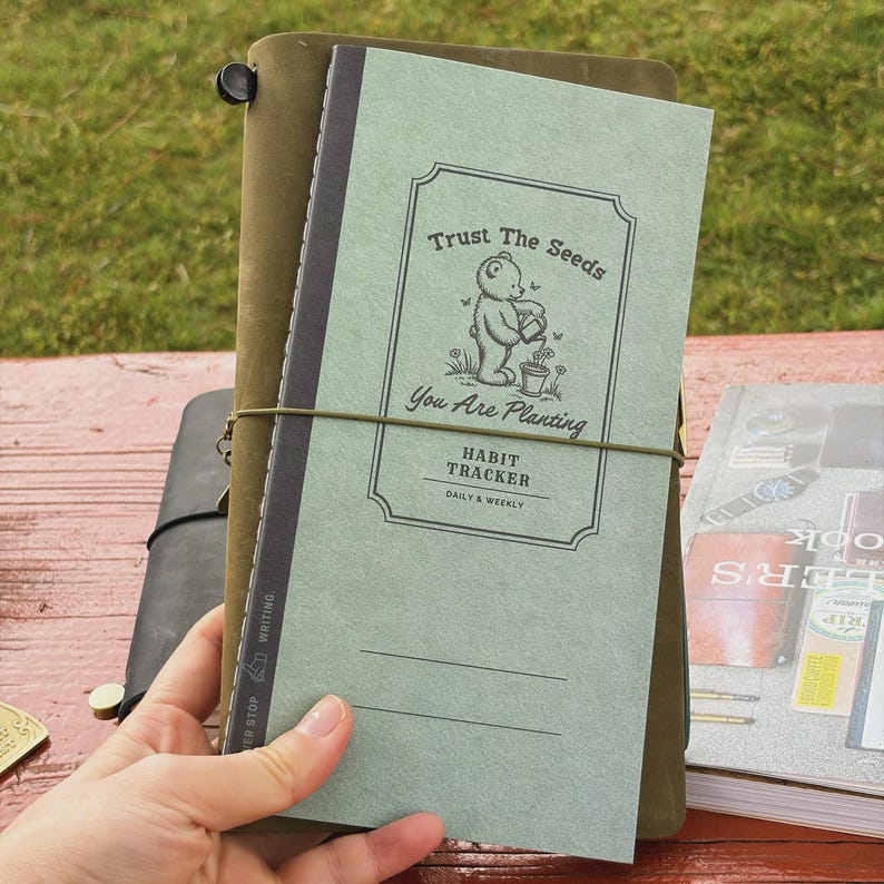 May include: A green habit tracker notebook with the text "Trust The Seeds" and "You Are Planting". The notebook is held together with an elastic band and is surrounded by other notebooks. The cover features a bear watering a plant.